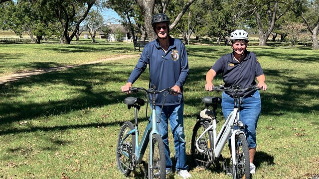 Two people in helmets stand next to bikes. In the background is a small, white house.