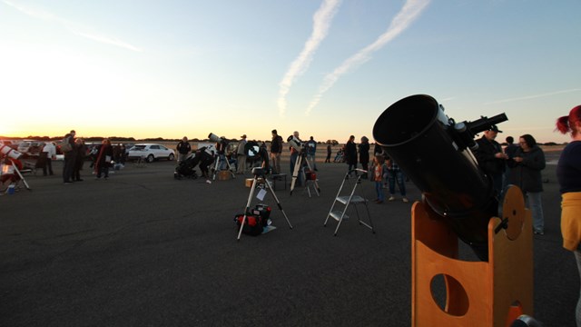 People gather around telescopes as the sun sets.