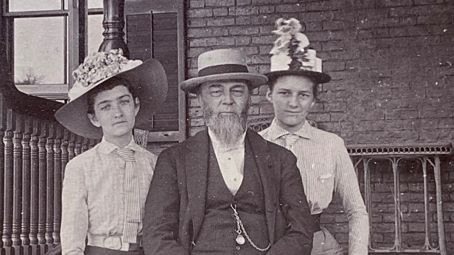 historic black and white photo of older man on sitting between two younger women on porch