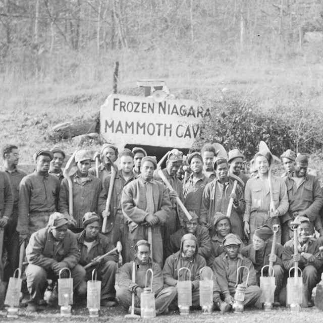 A black and white photo of a group of men posing for a group photo in front of a cave entrance. 