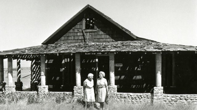 Black and white photo of two elderly woman talking in front of an old, vacant house.