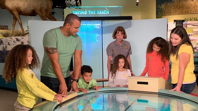 A uniformed ranger, two adults and four children look down at a colorful, circular table exhibit.