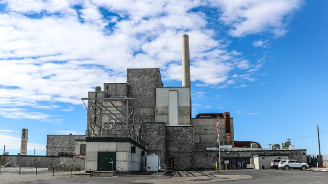 Multistory industrial grey concrete building with tall exhaust stack surrounded by paved lot