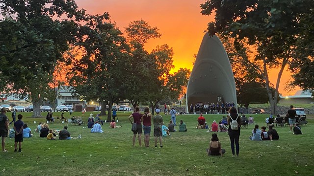 During sunset, a group of people on a large grassy area look toward stage with people. 