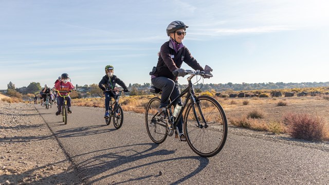  A woman and two young boys bicycle down a paved path next to bushes. The sky is blue