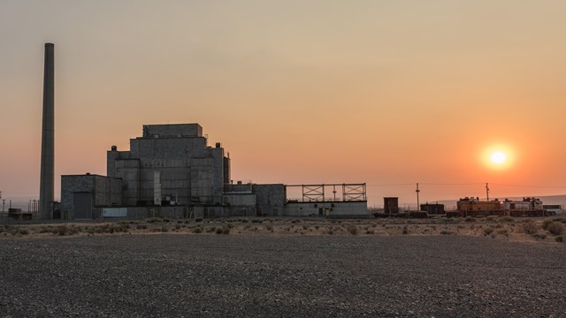 Orange sunset, a dark silhouette of large factory with single high smokestack.