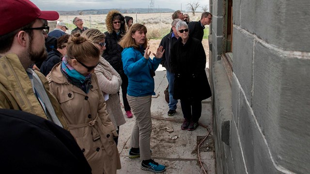 A group of people stand around a woman who is gesturing towards the gray wall of a building.