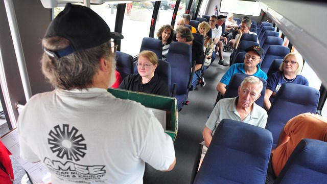 Several people seated on a bus while a guide stands speaking at the front of the bus.