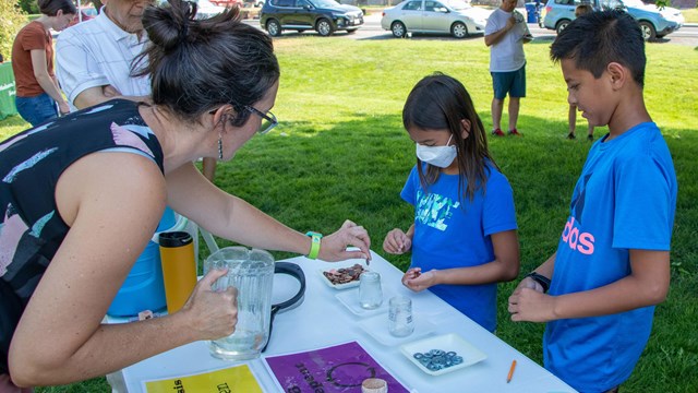 A woman holding a pitcher drops a penny in a glass of water on a table in front of 2 kids outside.