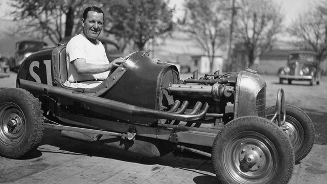 Black and white photo of old-time car with single seat and no roof, man in white shirt driving.