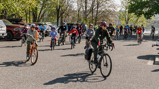 A group of 30 bicyclists are in a parking lot all riding in the same direction on a sunny morning. 