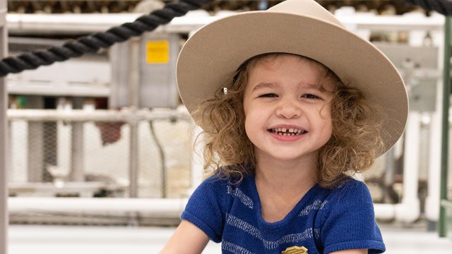 A young girl wearing a large campaign hat is seated on the floor smiling directly at the camera. 