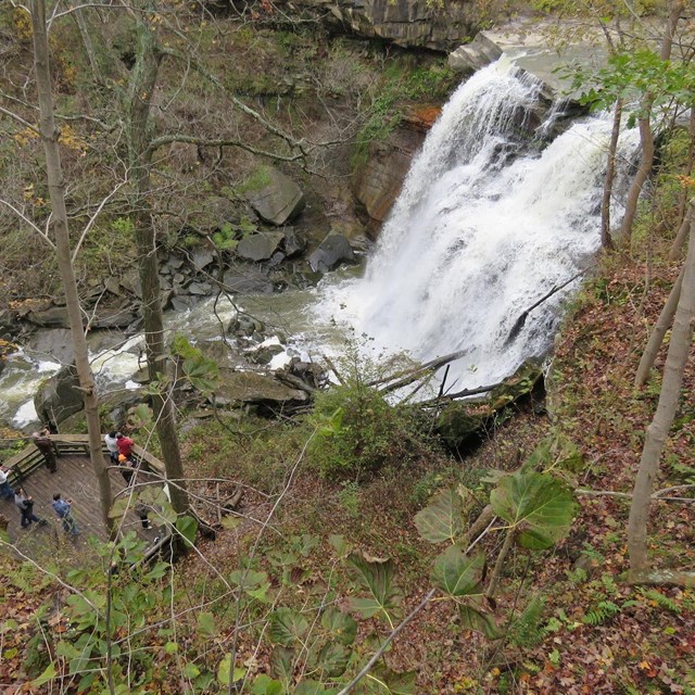 Viewed from above, people stand on a platform near frothy white water falling over a rocky ledge.