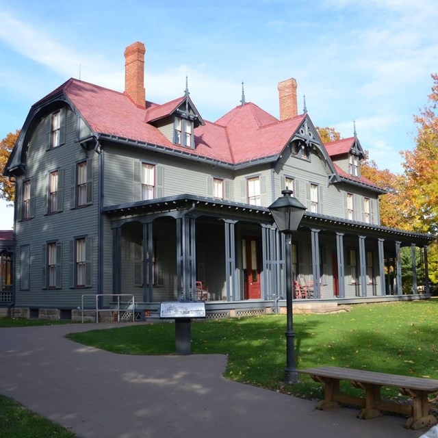 Large gray three-story house with a steep-sided red roof and large front porch. 