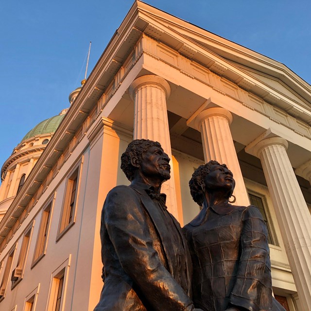 The statue of a Black man and woman under a large building with Greek-style columns. 