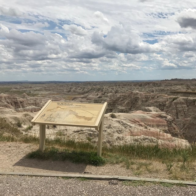 A wayside exhibit stands at the edge of drop off into badlands formations