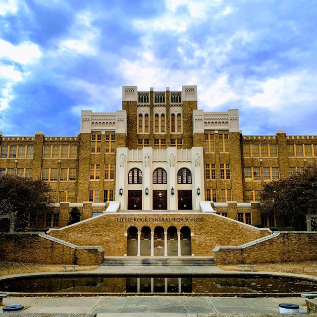 Front of a high school made of brown brick that rises to a high point in the middle with stairways.