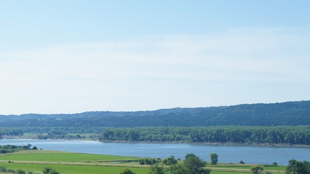 A fence and grassy field in the foreground with the river and bluffs in the background.