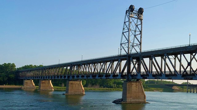 A metal suspension bridge stretches across the blue waters of the river.