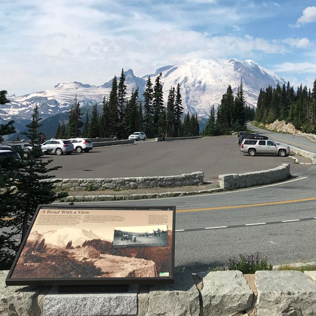 A wayside panel on a low rock wall overlooks a road leading towards a glaciated mountain peak.