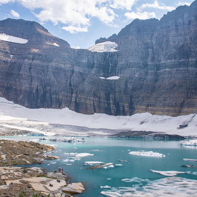 Steep mountains with glacier remnants and turquoise lake in foreground with floating chunks of ice.