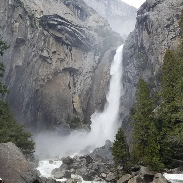 Waterfall gushes down granite cliff and conifer trees lining sides of rushing stream. 