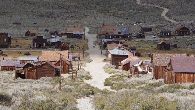 Wood frame buildings lining a dirt road in a desert setting. A road winds up the mountain in the bac