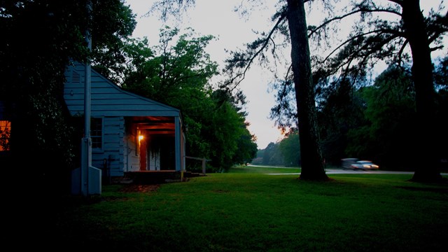 A dark photo of a cabin with a light showing through the window and lights on a truck on the road.