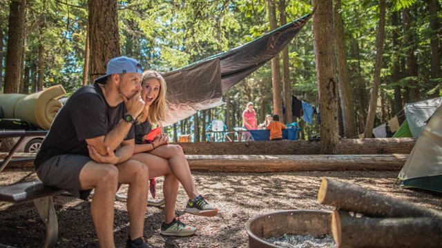 two people sit in a campsite in a forest with other campers in the background