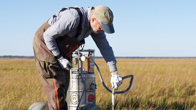 A field crew member bends down to stick a metal rod into a marsh