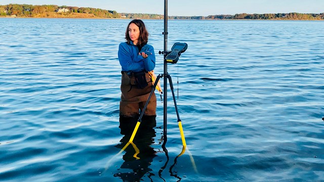 A field crew member stands in water and holds a tall tripod with a cylindrical device at the top