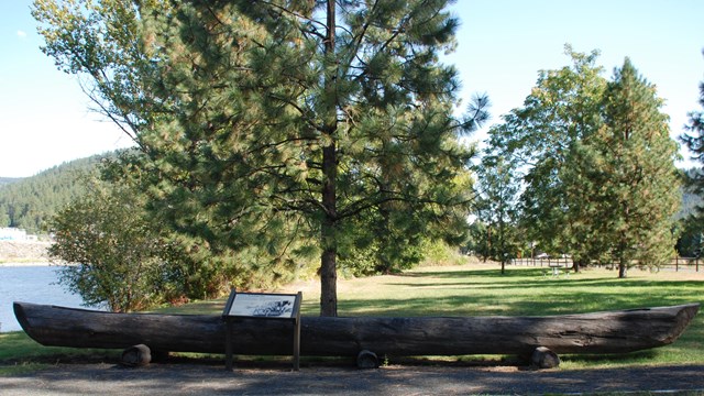 A wooden canoe sits in a grassy area next to a river.