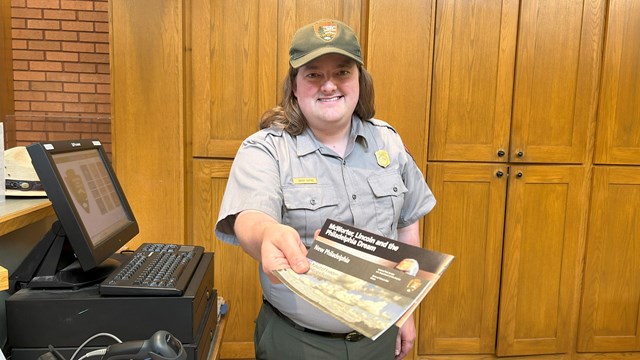 A park ranger smiles at you and reaches across a countertop to offer you two brochures.