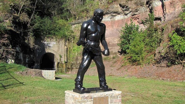 A black statue of a man holding a sledgehammer sits to the side in front of a railroad tunnel