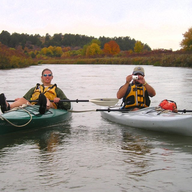 Two kayakers in their own boats rest on the calm waters of the river on a cloudy day.