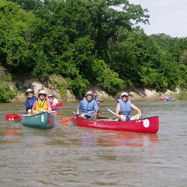 Two full canoes, red and green, each with two people, on a calm river.