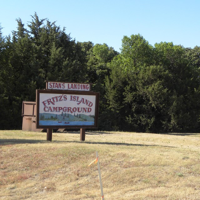 An old wood painted sign on the side of a gravel road with trees behind it.