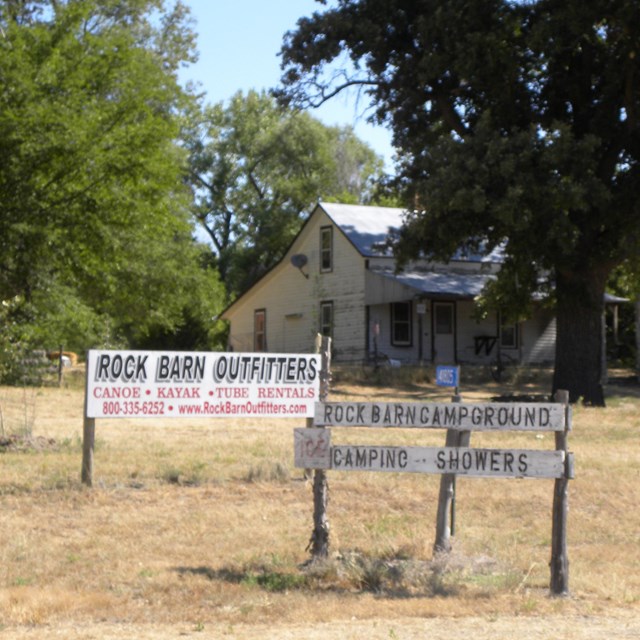 A wooden sign in a brown lawn with a building behind it through the trees