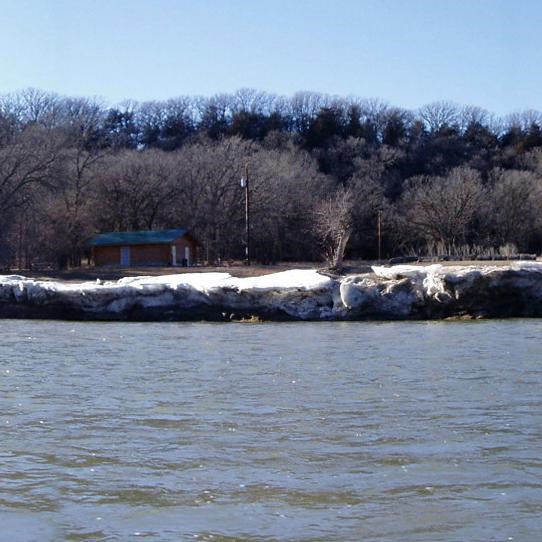 An Icy riverside with a bridge over the water and a building in the distance under the winter trees