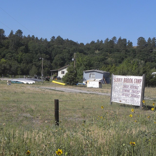 An old wooden sign in a grass field with canoes and kayaks on a trailer in the background