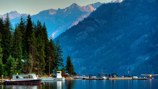 Boats and float plane docked along the shores of Lake Chelan with the mountains rising above. 