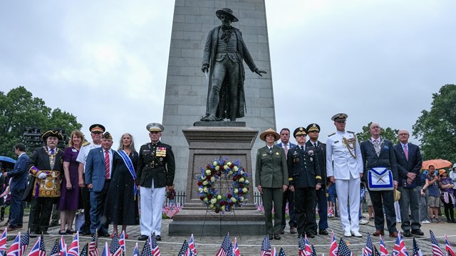 About a dozen people in uniforms in front of a statue and stone monument with American flags.