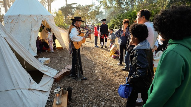 A person plays guitar in front of an audience next to two small canvas tents.