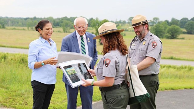 Four people, including two rangers in uniform, look at plans on a road next to a grassy battlefield.