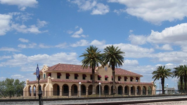 A long one story building with a covered walkway, set in the desert next to some palm trees.