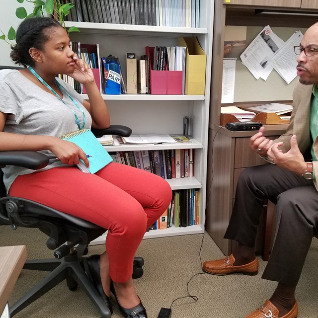A young woman with a notebook listens to a man speaking, seated beside each other in an office.
