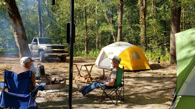 Two gentlemen next to a small fire ring at a campsite, tents and a truck are in the background