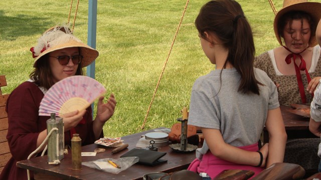 A young woman in a hat & dress era 1812 is seated behind a small table with period household items, 