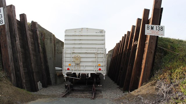 A boxcar rests on a rail track between two large wooden walls, backed by earth mounds that support t