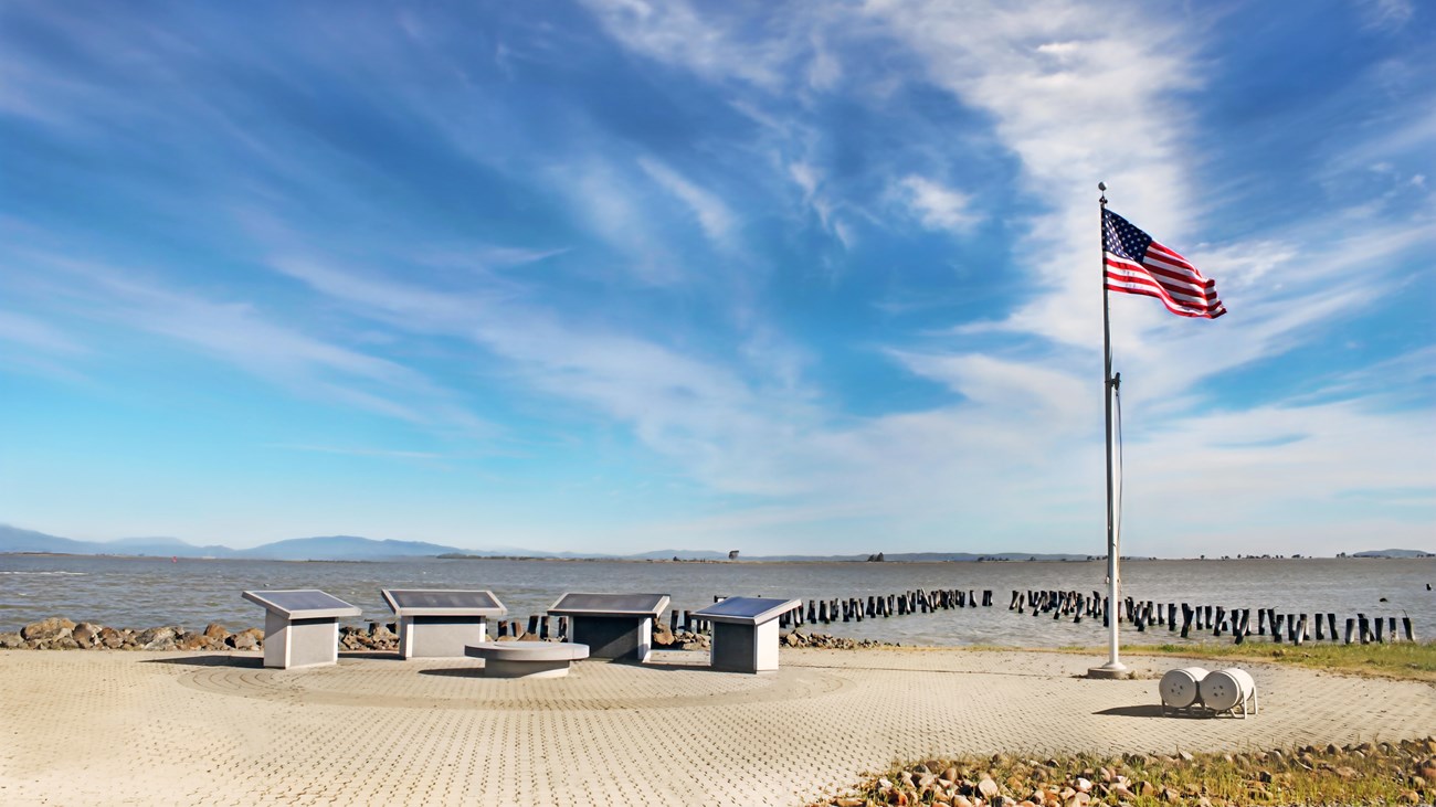 Four memorial stones sit on layered brick. An American flag waves in the wind. Bay water and hills. 